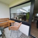 Terrasse mit Holzbank, Tisch und Rattan-Stuhl. Große Fensterfront zum Innenraum.