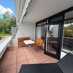 Terrasse mit Tisch, zwei Bänken und Liegeflächen. Blick auf grüne Umgebung und Parkplatz.