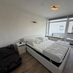Schlafzimmer mit Bett, Nachttisch und Sitzgelegenheit. Große Fensterfront mit Blick auf Balkon.