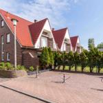Red tiled roof, brown facade, paved street with bike racks and green hedge.