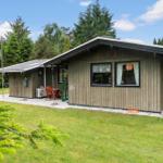 Holzhaus mit Terrasse, Garten und Bäumen. Fenster mit Vorhängen. Außentisch und Stühle.