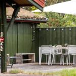 Terrasse mit Tisch, Stühlen und Sonnenschirm. Hintergrund: grüne Holzwand und Garten.