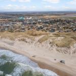 Strand mit Dünen und Küstenort im Hintergrund. Wellen brechen am Ufer.
