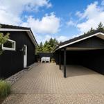 Schwarze Holzhütte mit Carport und gepflastertem Hof. Weiße Bank und Pflanzen im Garten.