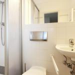 Badezimmer mit Dusche, Waschbecken und Toilette. Fliesenwand und Spiegel sichtbar.