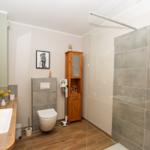 Badezimmer mit Dusche, Toilette und Waschbecken auf Holzunterlage.