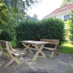 Holztisch und Bänke auf gepflastertem Platz vor grüner Hecke und Haus