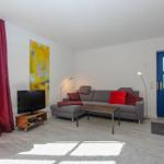 Wohnzimmer mit grauem Sofa, roten Kissen, blauer Tür und Fernseher auf Holzstand.