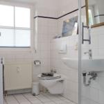Badezimmer mit Toilette, Waschbecken und Dusche. Weiße Fliesen und Fenster mit Jalousie.