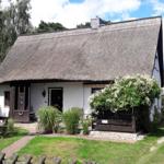 Ein Haus mit Strohdach, weißer Fassade und Garten. Holztor und Pflanzen sind sichtbar.