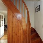 Holzgetreppe mit Geländer führt nach oben. Weiße Wände und Tür im Hintergrund.
