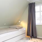 Ein Schlafzimmer mit Dachschrägen, weißem Bett, grauem Vorhang und Fenster.