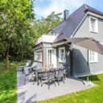 Graues Haus mit Terrasse, Tisch, Stühlen und Sonnenschirm. Umgeben von Grün und Bäumen.