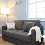 Ein grauer Sofa mit Kissen und Decke, Wand mit Schriftzug, Fenster und Beleuchtung.