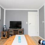 Ein Raum mit Holztisch, Fernseher, Tür und Fenster mit weißen Vorhängen.