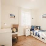 Schlafzimmer mit Bett, Schreibtisch und Fenster. Wandbilder und blaue Kissen.