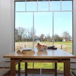 Holztisch mit Obstschale und Buch vor großem Fenster mit Blick auf Feld und Bäume.