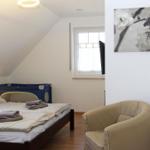 Schlafzimmer mit Bett, Sessel und Fenster. An der Wand hängt ein Bild mit Blumen.