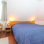 Schlafzimmer mit Bett, Nachttisch und Stuhl. Fenster mit Vorhängen und Bodenfliesen.