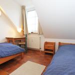 Zwei Betten, Schreibtisch, Fenster und Holzmöbel in einem Dachzimmer mit Terrakottboden.