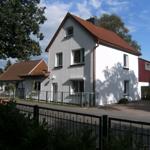 Weiße Villa mit rotem Dach und Metallzaun. Umgeben von Bäumen und Grünflächen.