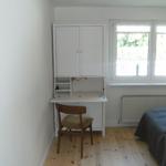 Schlafzimmer mit Schreibtisch, Stuhl und Fenster. Holzfußboden und Bett mit grauer Bettwäsche.