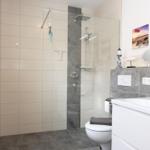 Bad mit Dusche, WC und Waschbecken. Fliesenwand und graue Bodenfliesen.