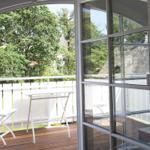 Durch die Glastür ist eine Terrasse mit Tisch und Stühlen zu sehen. Im Hintergrund sind Bäume und ein Dach sichtbar.
