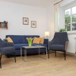 Ein Wohnzimmer mit blauem Sofa, zwei Sesseln und Holzmöbeln. An der Wand hängen Bilder.