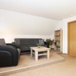 Wohnzimmer mit Sofa, Sessel, Tisch und Bücherregal. Holztür und Teppichboden.