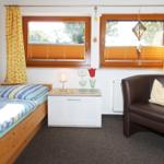 Ein Zimmer mit Bett, Sessel und zwei Fenstern mit Jalousien und Vorhängen.