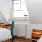 Zimmer mit Dachschrägen, zwei Betten, Holzschreibtisch und Fenster mit Vogeldekoration.