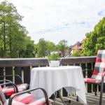 Terrasse mit Tisch, zwei Stühlen und Blumenkorb. Blick auf grüne Bäume und Häuser.