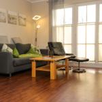 Wohnzimmer mit Sofa, Sessel, Tisch und Holzboden. Große Fenster mit Jalousien.