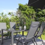 Terrasse mit Tisch, vier Stühlen und Sonnenschirm. Grün hinter dem Zaun.