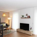 Wohnzimmer mit Sofa, Fernseher und Kaminofen. Fenster mit weißen Vorhängen.