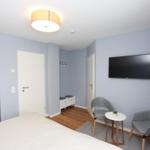 Schlafzimmer mit Bett, Fernseher und zwei Stühlen. Wandfarbe hellblau, Holzboden.
