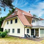 Ein zweistöckiges Haus mit rotem Dach und Terrasse. Garten und Sitzgelegenheiten sind vorhanden.