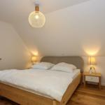 Schlafzimmer mit Doppelbett, Nachttisch und Fenster. Holzboden und Deckenleuchte.