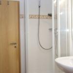 Badezimmer mit Dusche, Waschbecken und Spiegel. Holztür und weiße Fliesen.