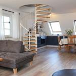 Wohnbereich mit Sofa, Küchenbereich und Tisch mit Stühlen. Eine Wendeltreppe führt nach oben.