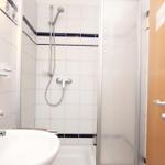 Badezimmer mit Dusche, Waschbecken und Spiegel. Tür führt in das Zimmer.