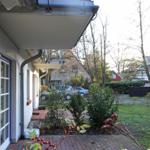 Vor dem Haus mit Balkon, Garten und Parkplatz. Bäume und Sträucher im Hintergrund.
