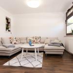 Wohnzimmer mit Ecksofa, runden Tisch und Holzboden. Fenster mit Jalousien und Pflanzen.