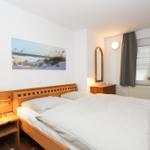 Ein Schlafzimmer mit Holzbett, Nachttisch und Fenster. An der Wand hängt ein Strandbild.