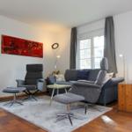 Wohnzimmer mit Sofa, zwei Sesseln, Tisch und Wandbild. Holzfußboden und Fenster mit Vorhängen.