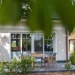 Haus mit Terrasse, Tisch und Stühlen. Grüne Pflanzen und Bäume umgeben das Gebäude.
