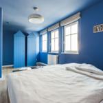 Schlafzimmer mit blauen Wänden, zwei Fenstern und einem Bett mit weißer Bettwäsche.