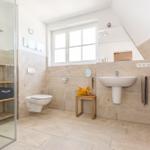 Badezimmer mit Dusche, Waschbecken und Toilette. Holzregale und Fenster sichtbar.