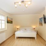 Schlafzimmer mit weißem Bett, Holzboden und Fenster. An den Wänden hängen Bilder und eine Wand-TV.